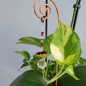 Könnte beinhalten: Ein Kristall-Sonnenfänger mit braunen und goldenen Perlen, die an einer Kupferspirale hängen. Der Sonnenfänger hängt an einer Pflanze.