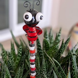 May include: A whimsical garden stake featuring a metal bee with a black head, large white eyes, and a red and black striped body. The stake is adorned with a decorative beaded stem in various colors, including red, black, and silver. The bee is set against a backdrop of green plants.