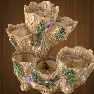 May include: A multi-tiered, tree-trunk-shaped planter with multiple openings for plants. The planter is painted in shades of brown and beige, with green moss and colorful flower and butterfly accents. A decorative item for indoor or outdoor use.