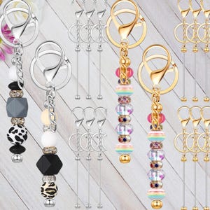 May include: A collection of keychains with decorative beaded charms. Some keychains have silver-colored hardware and charms with black, white, and gray beads. Other keychains have gold-colored hardware and rainbow-colored beads.