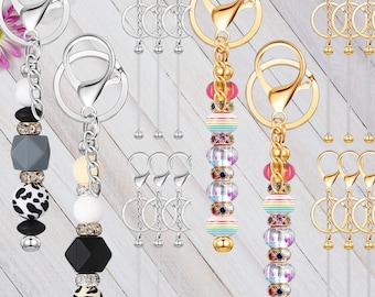 May include: A collection of keychains with decorative beaded charms. Some keychains have silver-colored hardware and charms with black, white, and gray beads. Other keychains have gold-colored hardware and rainbow-colored beads.