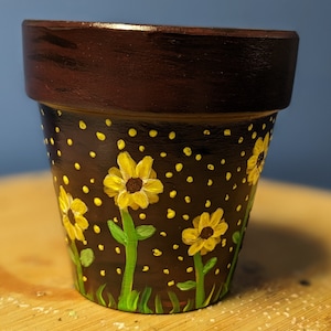 Può includere: Un piccolo vaso di terracotta dipinto di nero con pois bianchi e girasoli gialli.