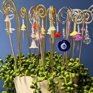 May include: A collection of decorative metal flower stakes with colorful flower charms and a blue evil eye charm. The stakes are arranged in a pot of green succulent plants.