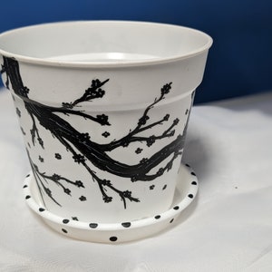 Peut inclure: Pot en céramique blanche avec un motif noir et blanc d'une branche d'arbre avec des fleurs. Le pot a une soucoupe assortie avec des pois noirs.