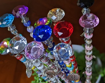May include: A collection of colorful crystal topped wands with beaded accents. The wands are made with various colors of beads and crystals, including pink, blue, green, red, and purple.