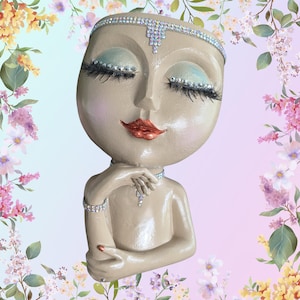 May include: A beige resin head planter featuring a stylized face with closed eyes, long black eyelashes, and metallic copper lips. The figure is adorned with clear rhinestone embellishments across the forehead, eyelids, and neck, and a bracelet.