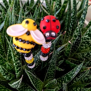 Puede incluir: Dos estacas decorativas para plantas, una abeja y una mariquita. La abeja tiene un cuerpo amarillo con detalles negros y alas blancas. La mariquita es roja con puntos negros. Ambas estacas tienen tallos enjoyados y están sobre plantas verdes.