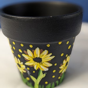 Op de afbeelding: Een kleine zwarte terracotta bloempot met een geschilderd ontwerp van gele zonnebloemen en gele stippen op een zwarte achtergrond.