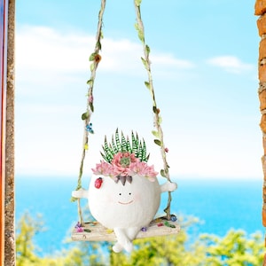 Puede incluir: Una maceta de cerámica blanca con forma de cara sonriente con brazos y piernas, colgada de una cuerda de columpio. La maceta está decorada con flores rosas y suculentas verdes. La maceta está colgada frente a un cielo azul y un océano.