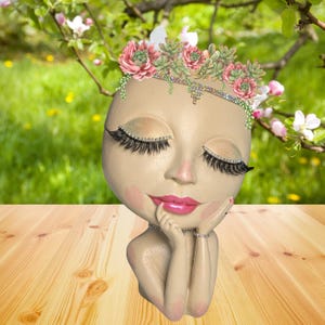 May include: A beige-colored face-shaped planter with closed eyes, long eyelashes, and pink lips. The planter is adorned with a floral crown of pink and green succulents and a jeweled headband. The figure rests its chin on its hands, with rings on the fingers.