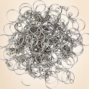May include: A collection of shiny, silver-colored key rings and chains, densely packed together. The key rings are round and vary in size, with some connected by small chains. The background is a soft, neutral beige.