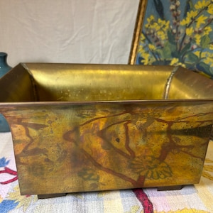 以下が含まれることがあります： 風化したパティナのある長方形の真鍮プランター。プランターはフレアトップで、4つの小さな足の上にあります。真鍮には赤茶色のアクセントがあります。背景には青いセラミックジャグがあります。