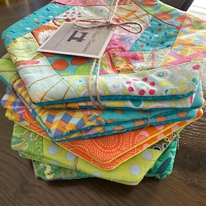 Puede incluir: Una pila de posavasos acolchados hexagonales coloridos, atados con hilo. Cada posavasos presenta un diseño de patchwork único con patrones y colores vibrantes, incluyendo azul, naranja, verde y amarillo. Una pequeña etiqueta con la letra "H" está adjunta.