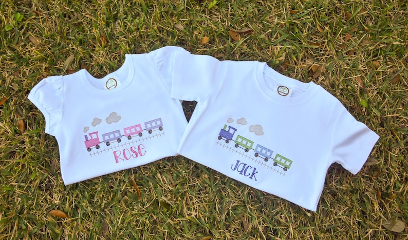 K&ouml;nnte beinhalten: Zwei wei&szlig;e T-Shirts mit einem Zugmotiv und den Namen "Rose" und "Jack", die darauf gestickt sind.