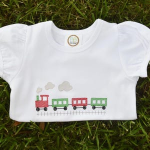 K&ouml;nnte beinhalten: Wei&szlig;es T-Shirt mit kurzen &Auml;rmeln und einem gestickten Design einer roten Lokomotive, die gr&uuml;ne G&uuml;terwagen zieht. Der Zug befindet sich auf einem Gleis mit Rauch aus dem Motor.