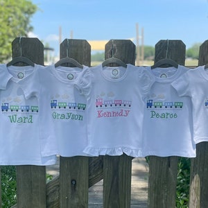 K&ouml;nnte beinhalten: Sechs wei&szlig;e T-Shirts mit gestickten Zugmotiven und Namen, darunter Weston, Ward, Grayson, Kennedy, Pearoe und Miller.