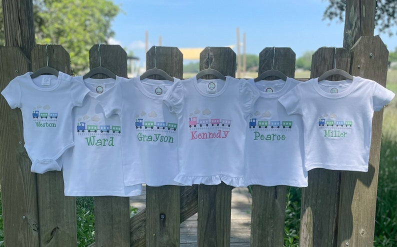 K&ouml;nnte beinhalten: Sechs wei&szlig;e T-Shirts mit gestickten Zugmotiven und Namen, darunter Weston, Ward, Grayson, Kennedy, Pearoe und Miller.