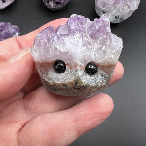 May include: A purple amethyst crystal carved into the shape of a hedgehog with black eyes. The crystal is rough and has a natural, organic look.