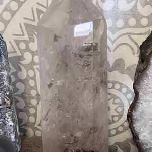 May include: A large, clear quartz crystal point with a natural, rough base. The crystal is translucent and has a slight smoky hue. It is approximately 10 inches tall.