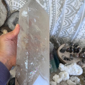 May include: A large, clear quartz crystal point with iridescent rainbow inclusions. The crystal is approximately 10 inches tall and has a smooth, polished surface.
