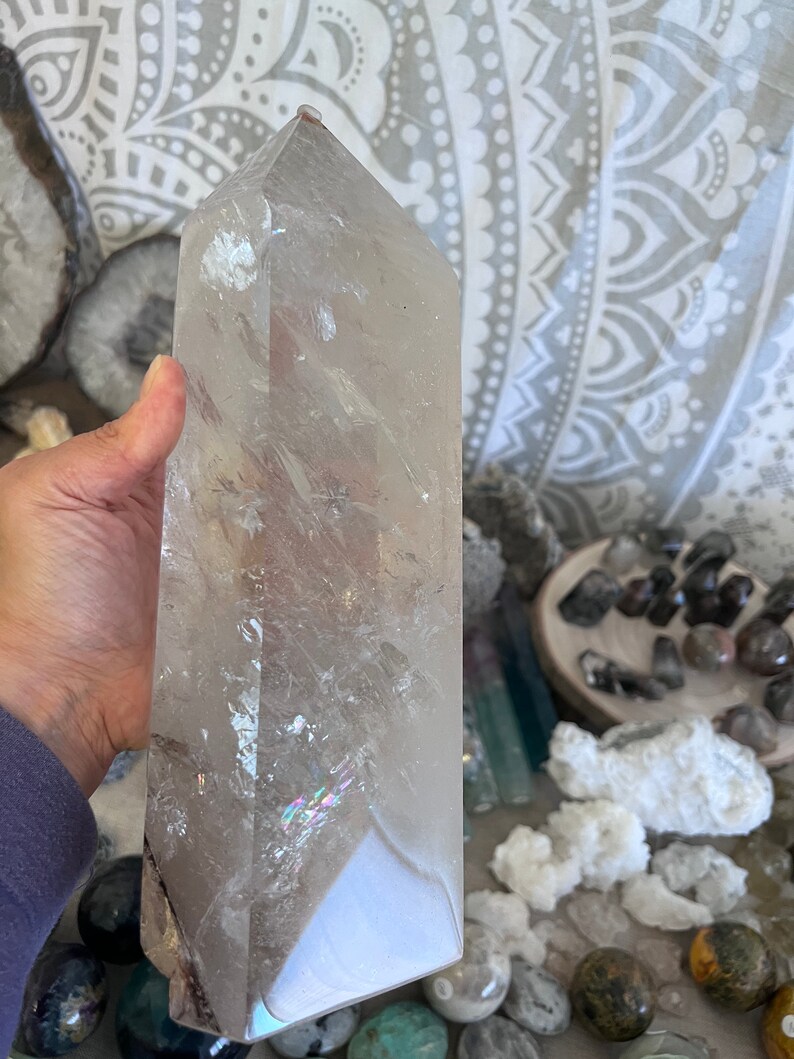 May include: A large, clear quartz crystal point with iridescent rainbow inclusions. The crystal is approximately 10 inches tall and has a smooth, polished surface.