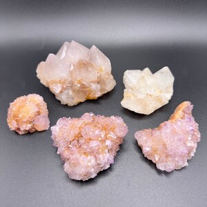 May include: Five natural crystal clusters in shades of beige, peach, and lavender are arranged on a dark gray surface. The crystals vary in size and shape, showcasing a range of textures and formations, ideal for collectors.