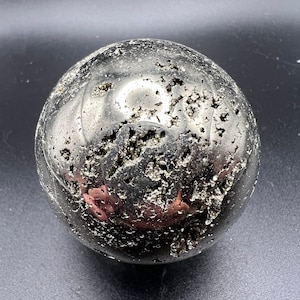 May include: A smooth, round, silver pyrite sphere with a textured surface. The sphere is made of natural stone and has a metallic sheen.