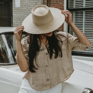 May include: A woman wearing a wide-brimmed straw hat with a tan linen shirt and white pants. The hat has a flat crown and a wide brim.