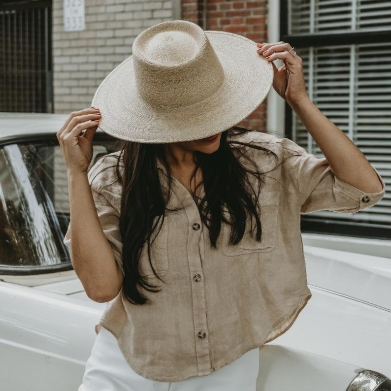 May include: A woman wearing a wide-brimmed straw hat with a tan linen shirt and white pants. The hat has a flat crown and a wide brim.