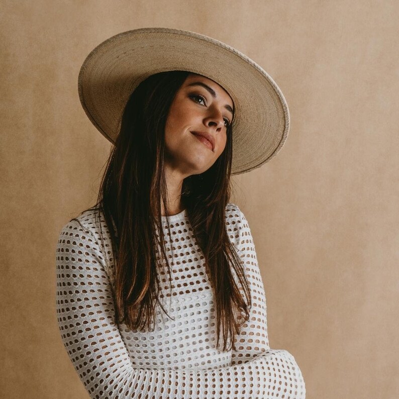 May include: A woman wearing a white, crocheted, long-sleeved shirt with a wide-brimmed, light brown straw hat.