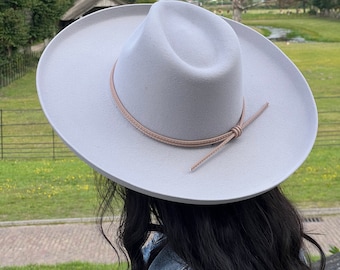 May include: A light gray felt fedora hat with a light brown leather band and a tied bow. The hat is worn by a person with long dark hair.