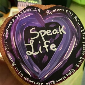 May include: A heart-shaped wooden plaque with a painted purple heart design. The words "Speak Life" are in white script in the center. Various Bible verses are written around the heart's edge in white. The plaque has a natural wood grain edge.