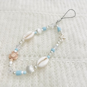 Puede incluir: Un dije de teléfono con cuentas con un tema de playa. El dije incluye conchas blancas, una estrella de mar y un dije de tortuga, intercalados con perlas blancas, cuentas transparentes y cuentas azul claro. Se adjunta un lazo negro.