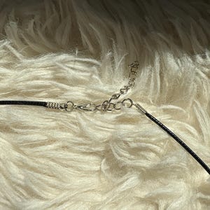 K&ouml;nnte beinhalten: Eine schwarze Halskette mit einer silbernen Kette und einem Verschluss. Die Halskette liegt auf einer flauschigen, wei&szlig;en Oberfl&auml;che. Die Kette ist mit einem schwarzen Band verbunden.