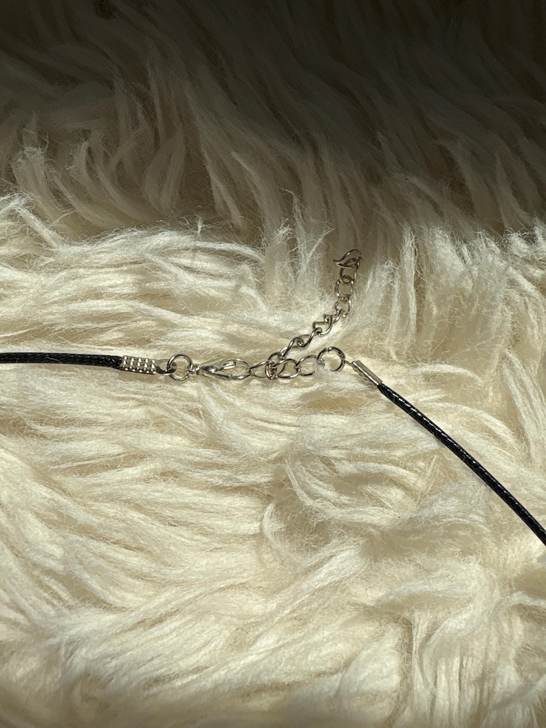 K&ouml;nnte beinhalten: Eine schwarze Halskette mit einer silbernen Kette und einem Verschluss. Die Halskette liegt auf einer flauschigen, wei&szlig;en Oberfl&auml;che. Die Kette ist mit einem schwarzen Band verbunden.