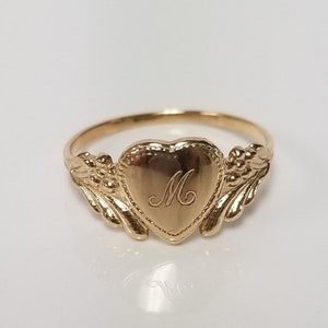 May include: A gold heart-shaped ring with a delicate floral design on the sides. The ring has an engraved letter "M" on the heart.