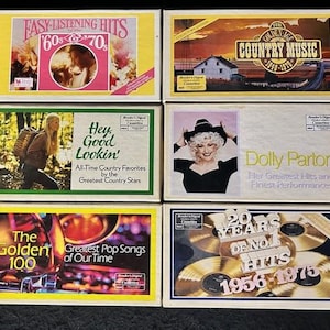 Könnte beinhalten: Eine Sammlung von sechs Kassetten mit verschiedenen Country- und Pop-Musikthemen. Die Kassetten zeigen Künstler wie Dolly Parton und die Golden 100. Die Kassetten befinden sich in einer weißen Pappbox mit einem farbenfrohen Design.