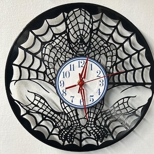 Puede incluir: Reloj de vinilo negro con una silueta de Spider-Man en un diseño de telaraña. La esfera del reloj es blanca con detalles azules y números negros.
