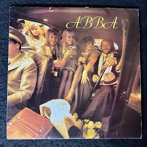 May include: A square vinyl record cover featuring the band ABBA inside a car. Four people are seated, holding glasses and a bottle. The text ABBA is printed in gold in the upper right corner. The scene is dimly lit with warm interior lighting.