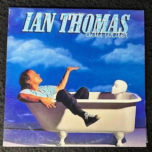 Könnte beinhalten: Albumcover für "Ian Thomas - Add Water". Das Bild zeigt einen Mann in einer Badewanne mit einem Schaufensterpuppenkopf vor einem blauen Himmel mit Wolken. Der Albumtitel und der Künstlername sind in blauer Schrift.