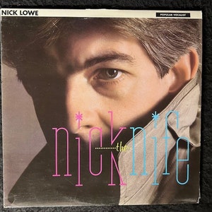May include: A vintage record album cover featuring a close-up of Nick Lowe's face. The album title "Nick the Knife" is written in pink and blue letters with a star on either side of the title.