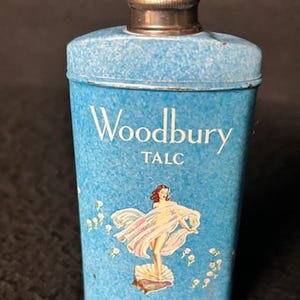 May include: A blue metal tin with a silver lid. The tin has a vintage design with the words "Woodbury TALC" and a woman in a white dress standing on a seashell.