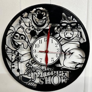 Pode incluir: Relógio de disco de vinil preto com personagens do The Muppet Show. O mostrador do relógio é branco com números pretos e ponteiros vermelhos. O logotipo do The Muppet Show está na parte inferior. O relógio está montado em uma parede branca.