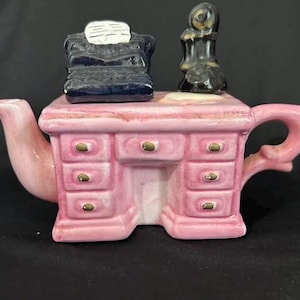 May include: A ceramic teapot shaped like a pink office desk with six drawers featuring gold-coloured knobs. On top of the desk sits a dark blue typewriter with a white paper insert and a black telephone receiver. The teapot has a curved spout and handle.