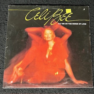 May include: A vinyl record cover featuring a woman in a red outfit with the title "Celli Bee Fly Me on the Wings of Love" in yellow text.
