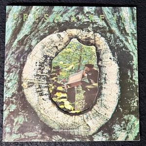 Op de afbeelding: Een vintage vinyl platenhoes met de tekst "FRESH AIR III" in grote, vetgedrukte letters. De hoesillustratie toont een close-up van een boomstam met een rond gat dat een scène van twee mensen in een bosomgeving onthult. De tekst "FRESH AIR III BY MANHATTAN TRANSFER" is zichtbaar onderaan de afbeelding.