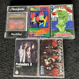 May include: A collection of eight cassette tapes featuring various artists, including Headpins, The Rolling Stones,  Warrant, The Pretenders, and KISS. The tapes are in clear plastic cases with colourful artwork and band names printed on the front.