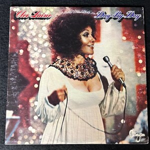 Può includere: Una copertina di un disco in vinile con Cleo Laine, una cantante, che tiene un microfono. La copertina è rossa, bianca e dorata con uno sfondo scintillante. Il titolo dell'album è "Day By Day".