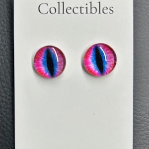 May include: A pair of round stud earrings with a cat's eye design. The earrings have a pink and blue iris and a black pupil. The card reads "J & S Vintage Collectibles" and "www.jandsvintagecollectibles.com".