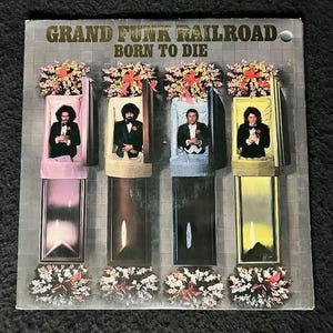 May include: A vinyl record album cover featuring the band Grand Funk Railroad. The cover art depicts four men in suits lying in coffins with flowers on top. The album title "Born to Die" is printed in gold lettering.